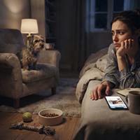 Mujer triste descansando en el sofá de su casa junto a su perro por la noche, mirando su teléfono móvil en una sala de estar tenuemente iluminada.